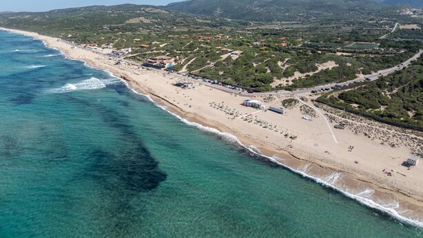 Vista aerea di una zona costiera con spiaggia, acque blu cristalline e terreno collinare sullo sfondo.