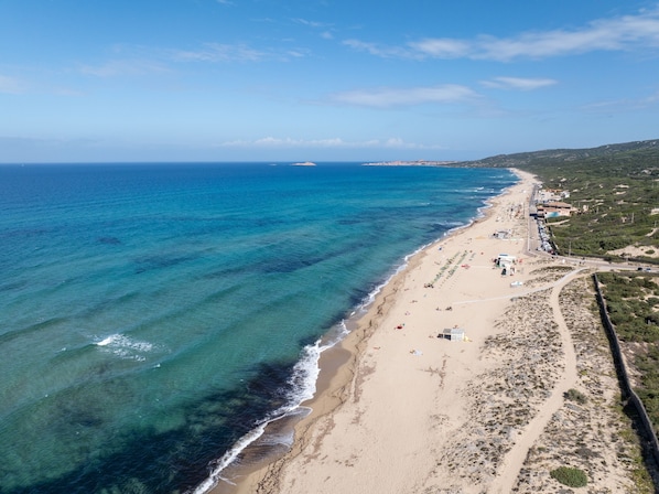 An aerial view of a coastal beach with clear blue waters, sandy shore, and a few buildings along the coastline. - Comfy Flat With sea View (Badesi)