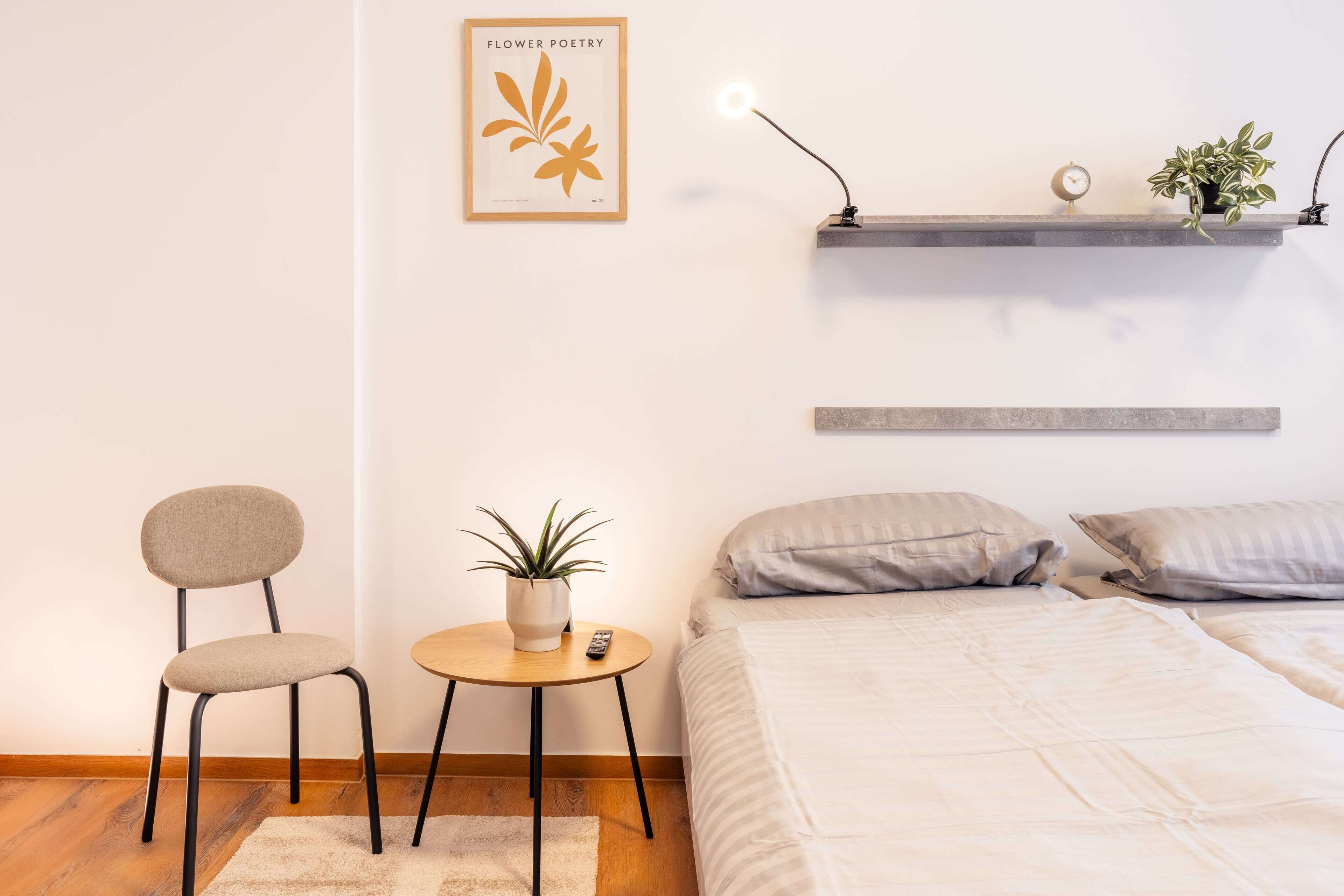 Un dormitorio con una cama, una silla, una mesita con una planta, un estante de pared con un reloj y otra planta, y un cuadro enmarcado en la pared.
