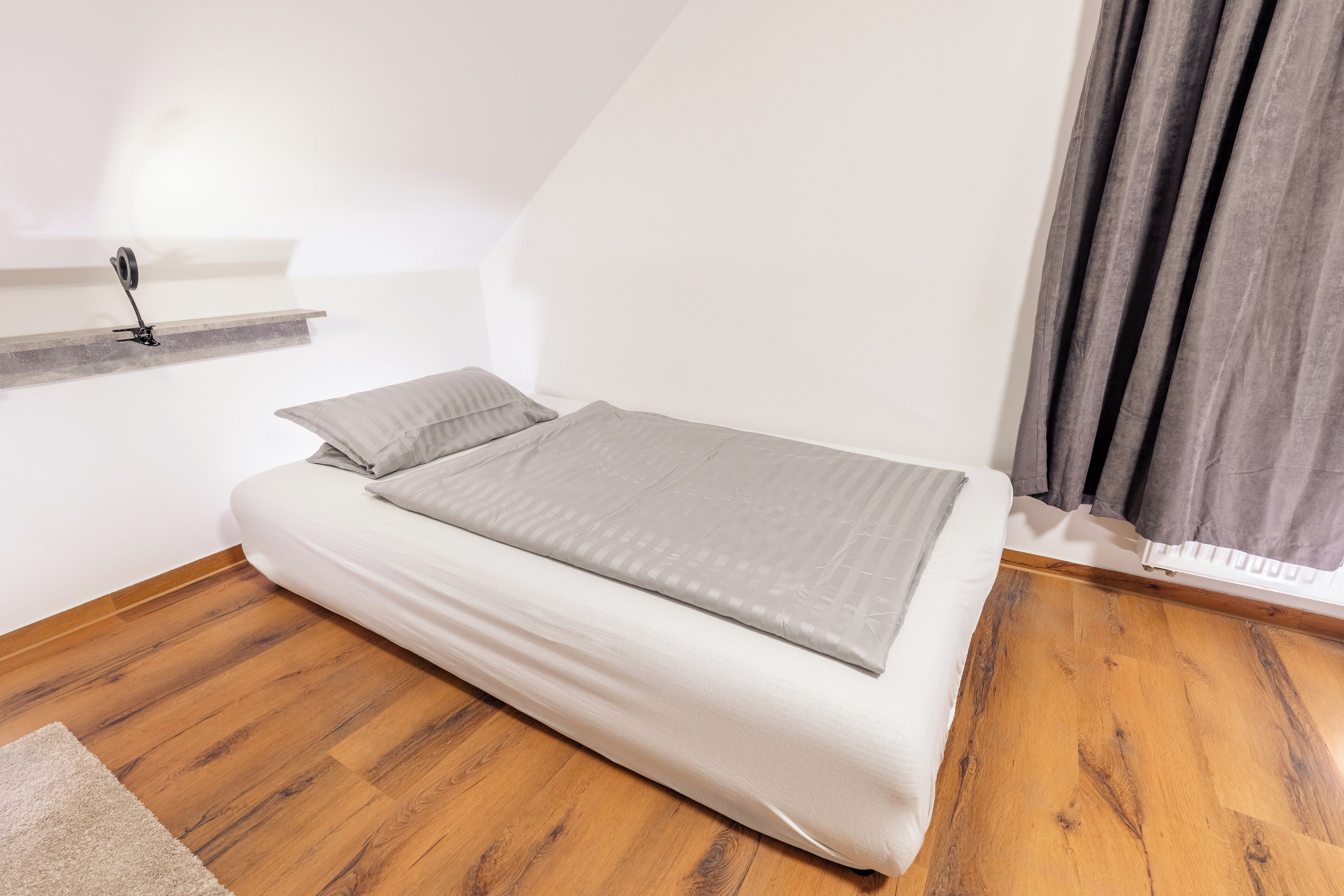 A small, neatly made bed with a grey blanket in a room with wooden flooring and a white wall.