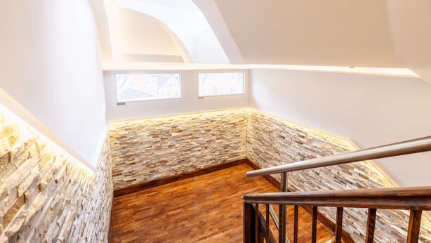 Un escalier avec un mur en pierre et un plancher en bois, menant à une pièce dotée d’un plafond en pente et d’une fenêtre.
