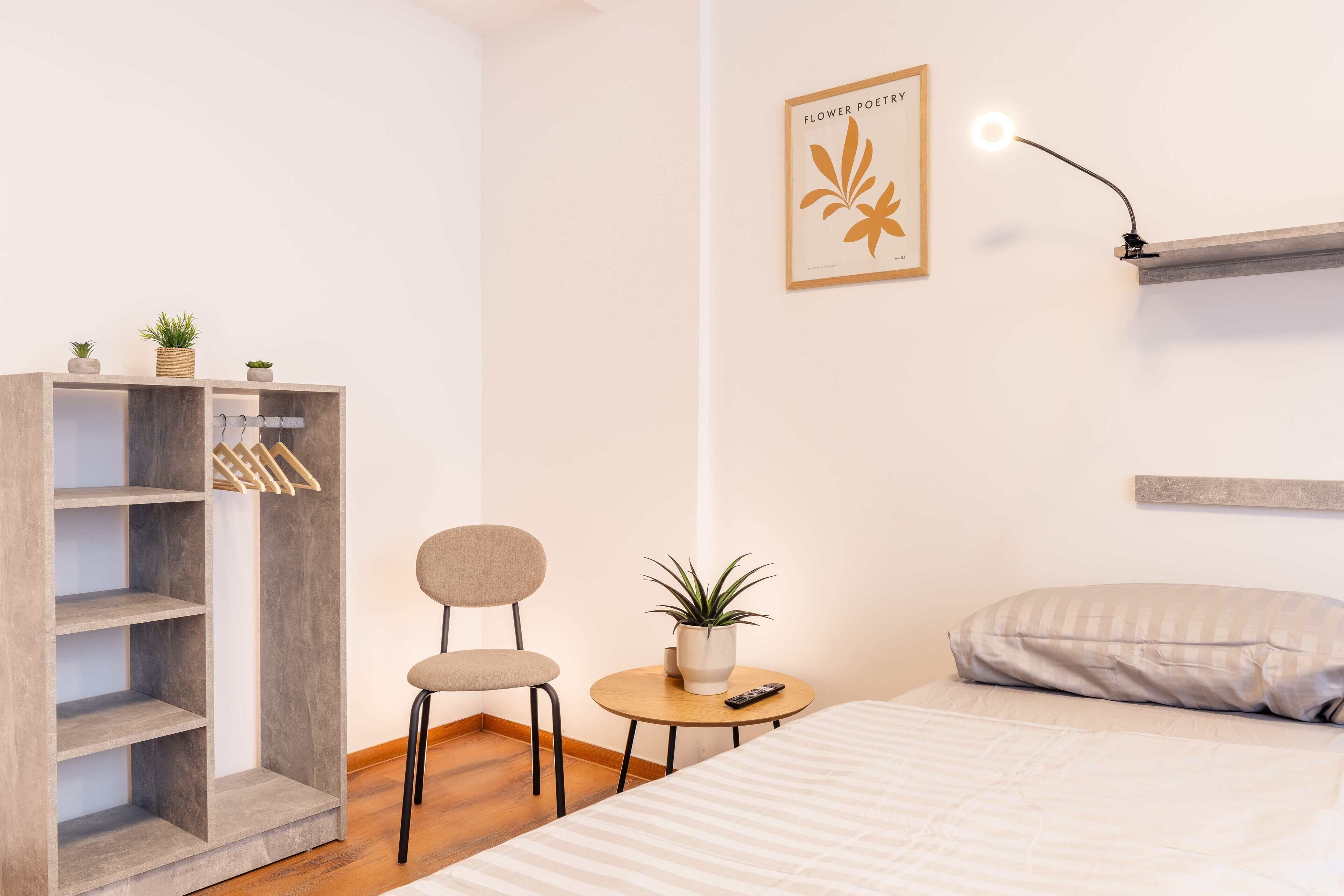 A bedroom with a bed, a small round table with a plant, a chair, a wardrobe, and a wall-mounted light fixture.
