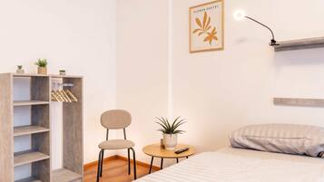 A bedroom with a bed, a small round table with a plant, a chair, a wardrobe, and a wall-mounted light fixture.