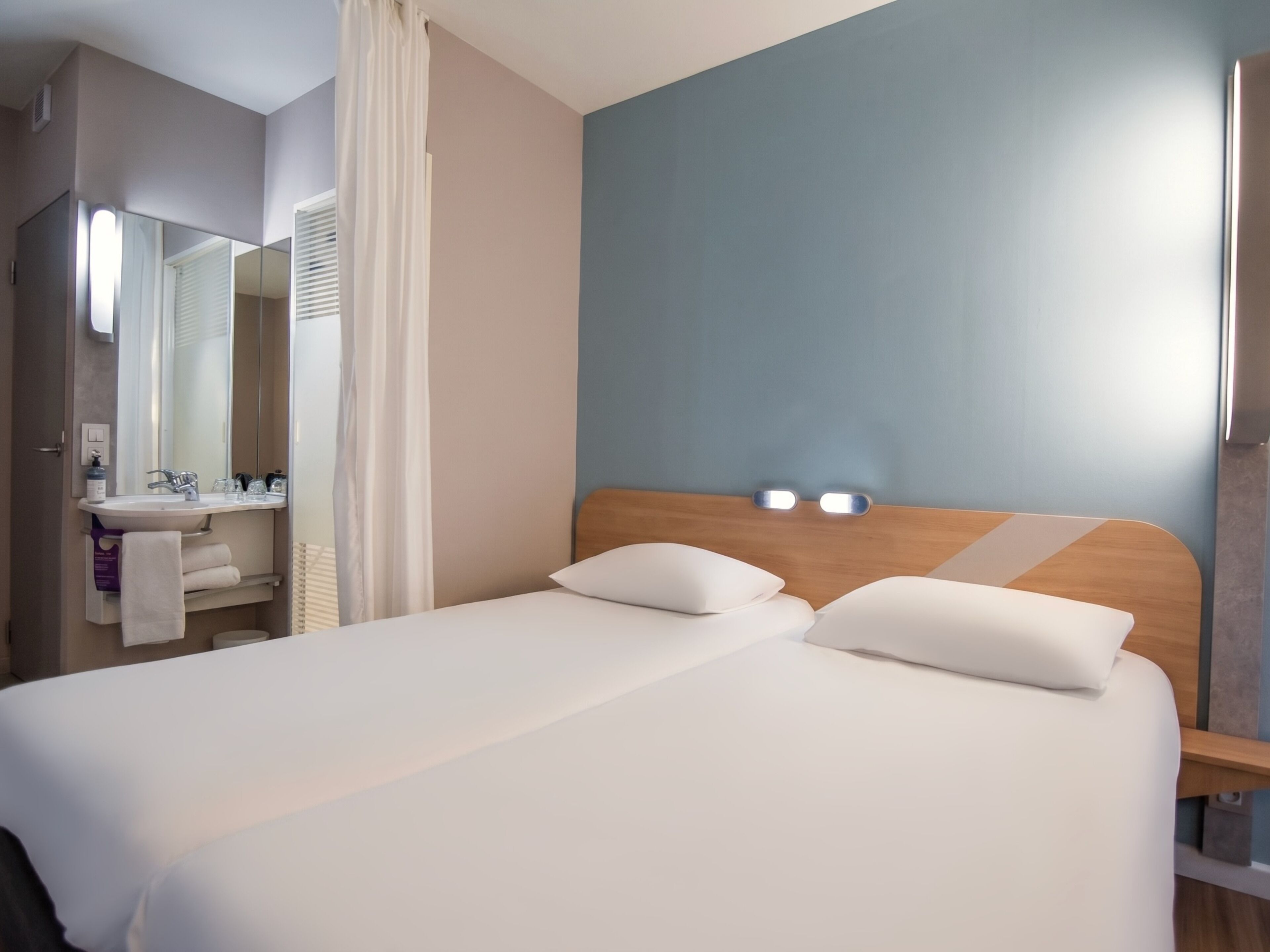 A hotel room with a bed, a bedside table, a mirror, a sink, and a towel rack.