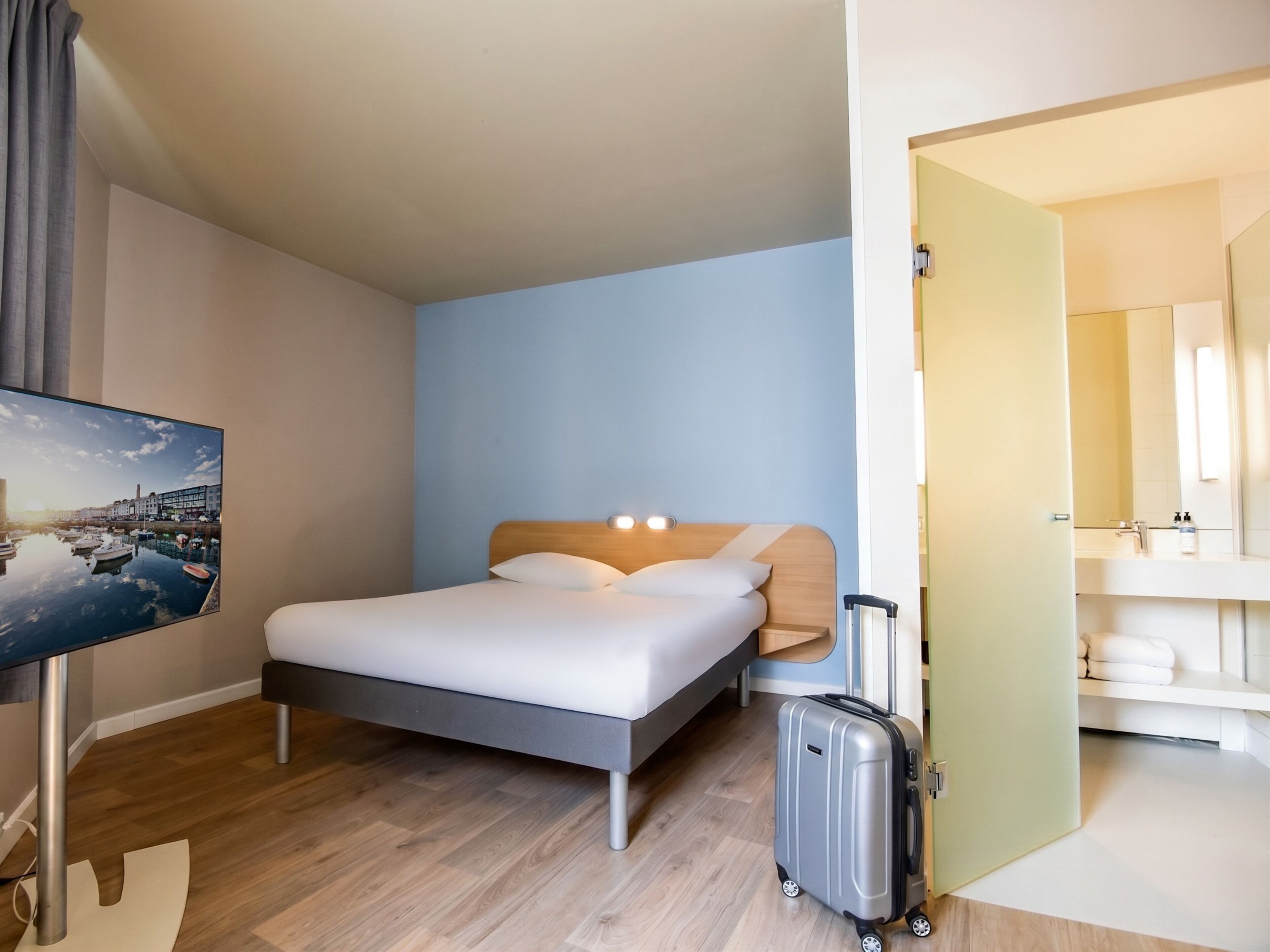 Habitación de hotel con cama, maleta, televisión y baño visible a través de una puerta abierta.