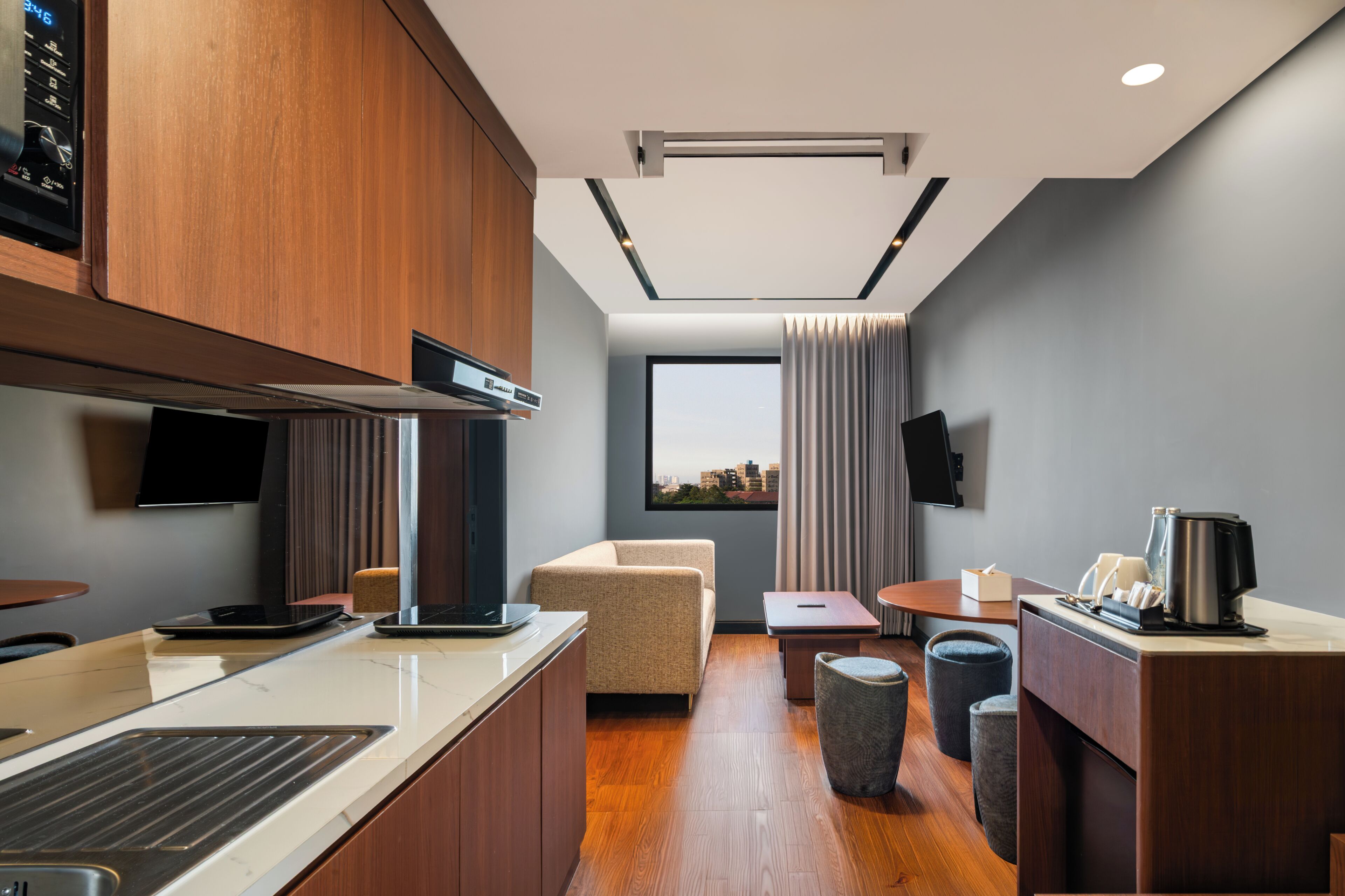 A modern hotel room with a kitchenette, a seating area with a sofa and a small table, a window with a view of the city, and a flat-screen TV mounted on the wall.