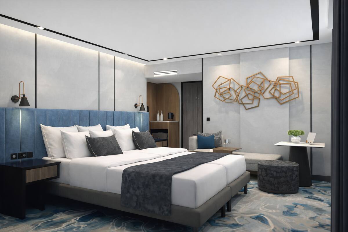 A modern hotel room with a large bed, a bedside table, a nightstand, a small seating area, and a decorative wall piece.
