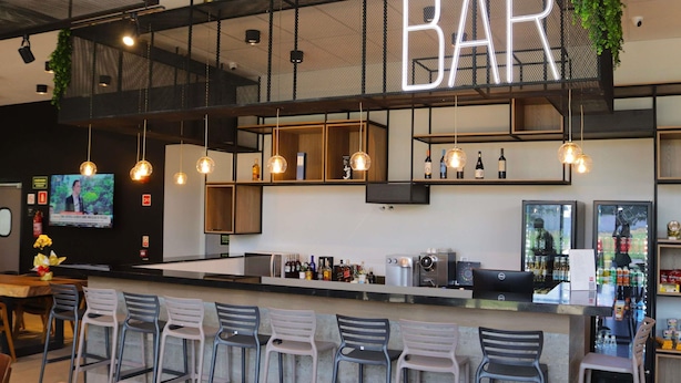 A bar area with a neon sign, hanging lights, bar stools, a counter with a coffee machine, and shelves with bottles. - ibis Primavera Do Leste (Primavera do Leste)