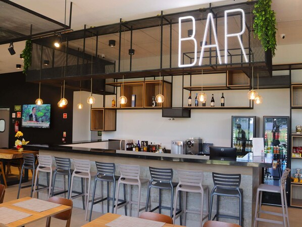 Zona de bar con un letrero de neón, luces colgantes, taburetes, una barra con una máquina de café y estantes con botellas.