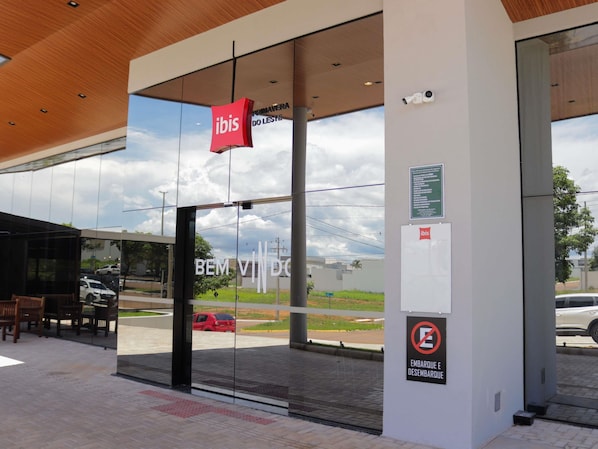 A modern building entrance with a glass facade, a red sign with 'ibis' logo, and a no parking sign. - ibis Primavera Do Leste (Primavera do Leste)