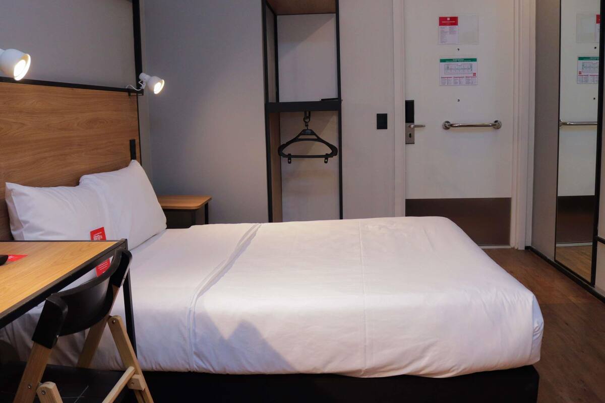 A hotel room with a bed, a desk, a chair, a wardrobe, and a door with a mirror.
