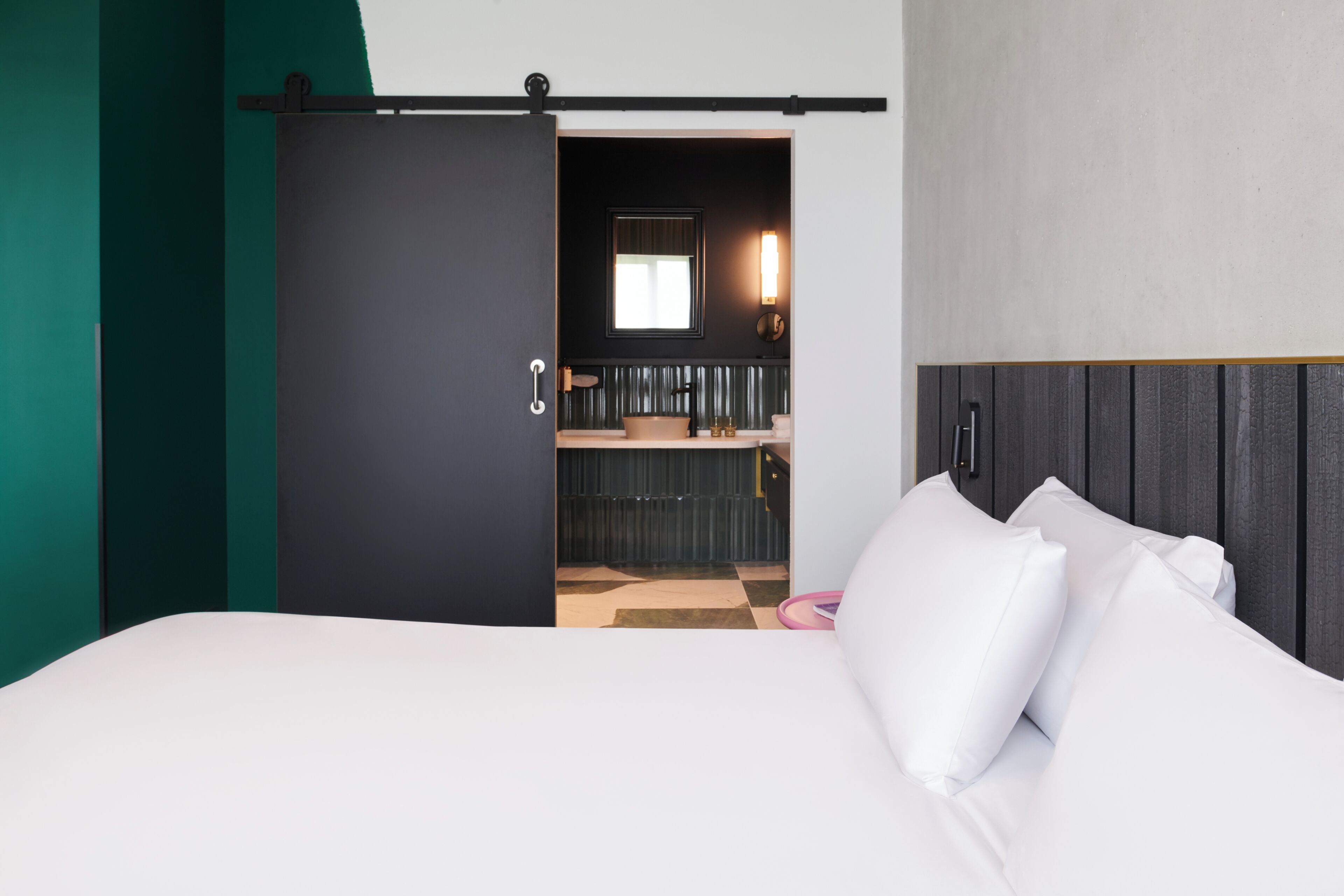 A hotel room with a bed, a sliding door to a bathroom, a checkered floor, and a window with a light fixture.