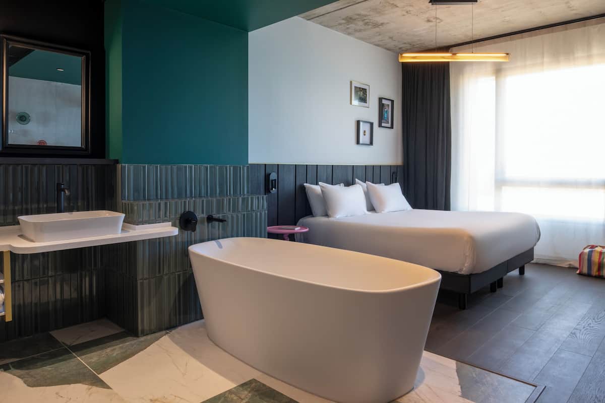 Habitación de hotel moderna con una bañera grande, una cama doble, un tocador con lavabo y un amplio ventanal con cortinas.