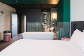 Une chambre d’hôtel moderne avec une grande baignoire blanche, un plancher en bois, un mur d’accent vert et une zone de douche bien visible.