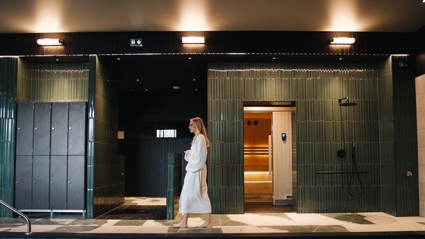 A woman in a white robe walking towards a room with a shower and a towel rack. - Mercure Quimper Kerlic (Quimper)