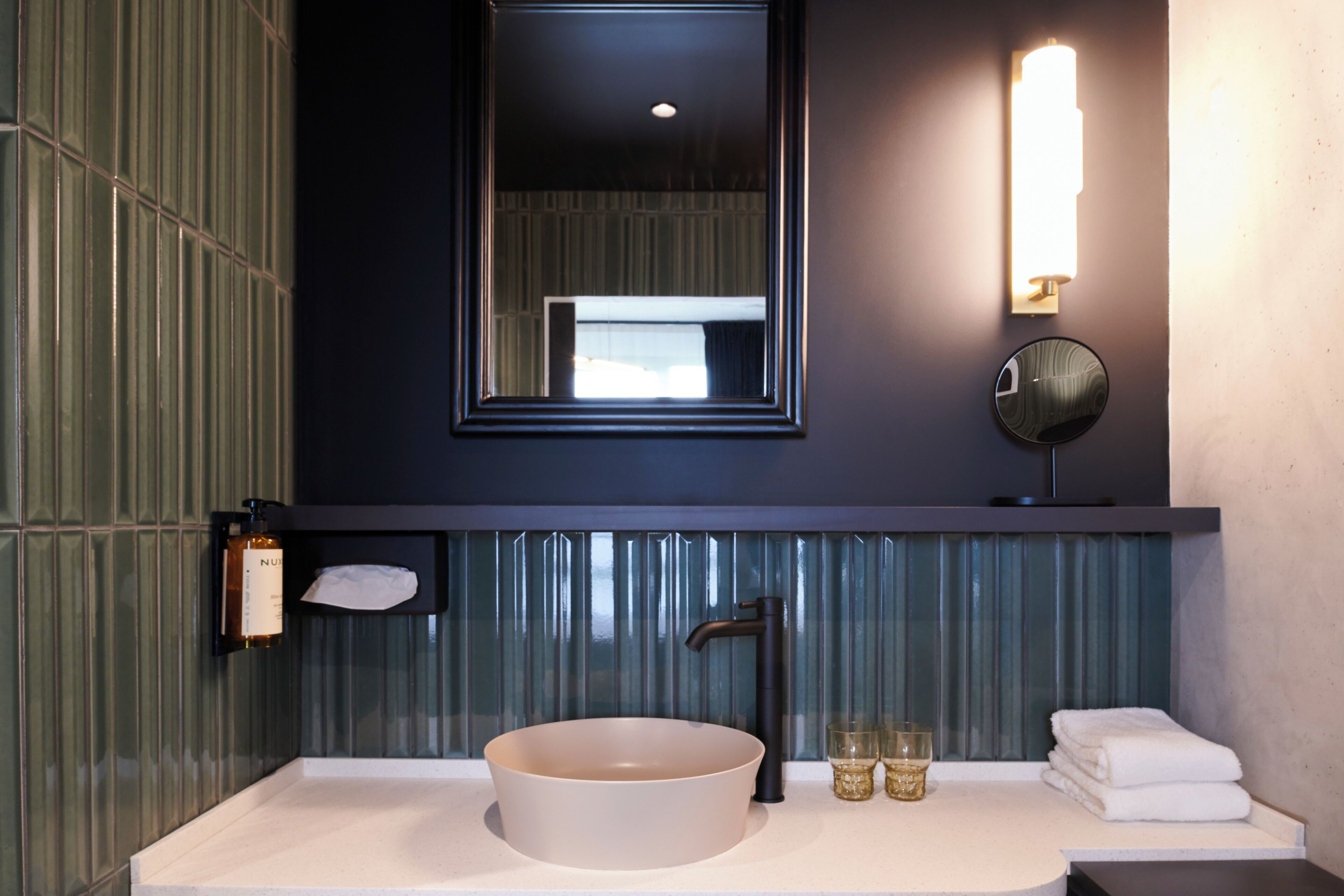 A modern bathroom with a round sink, a mirror, a towel rack, and a bottle of hand soap.