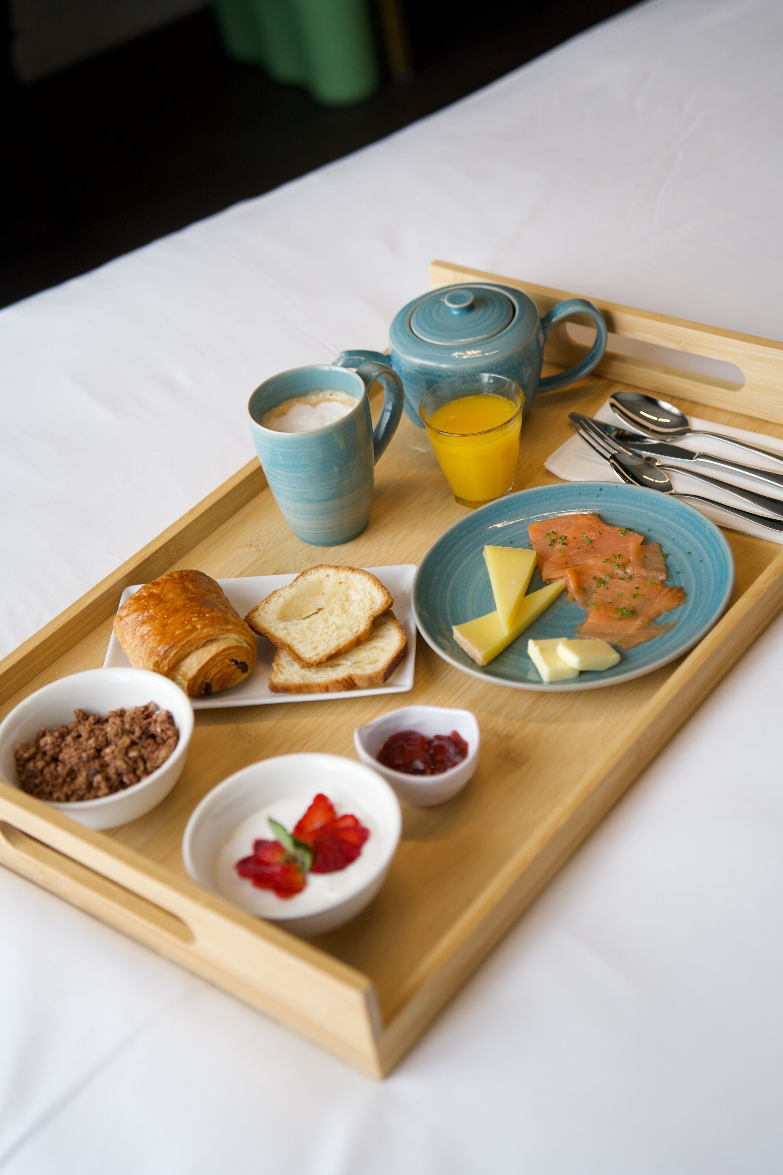 Bandeja de desayuno con cruasanes, tostadas, salmón ahumado, queso, mantequilla, una tetera, un vaso de jugo de naranja y recipientes pequeños de mermelada y yogur.