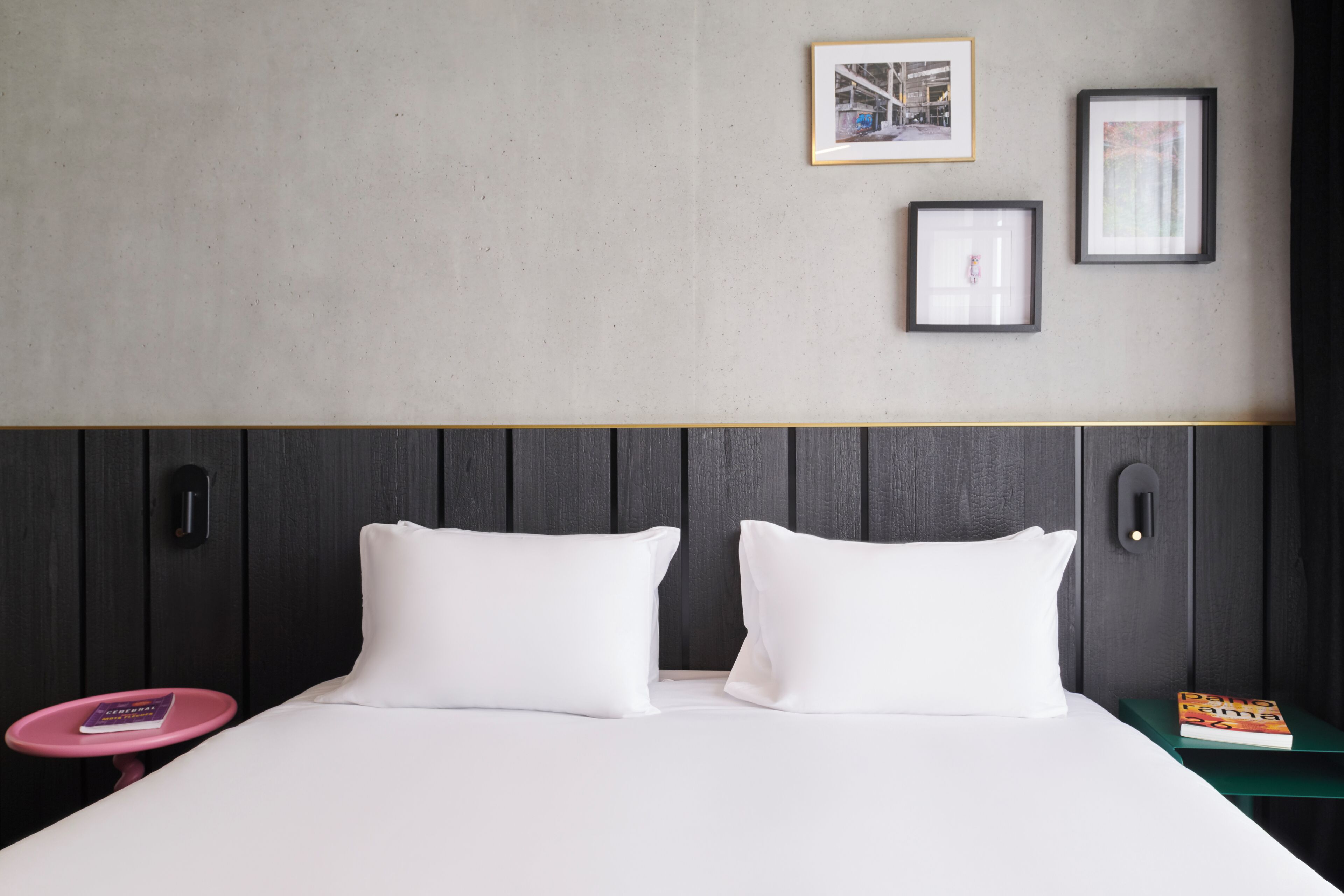 A hotel room with a bed, two pillows, a small pink table, a green nightstand, and two framed pictures on the wall.