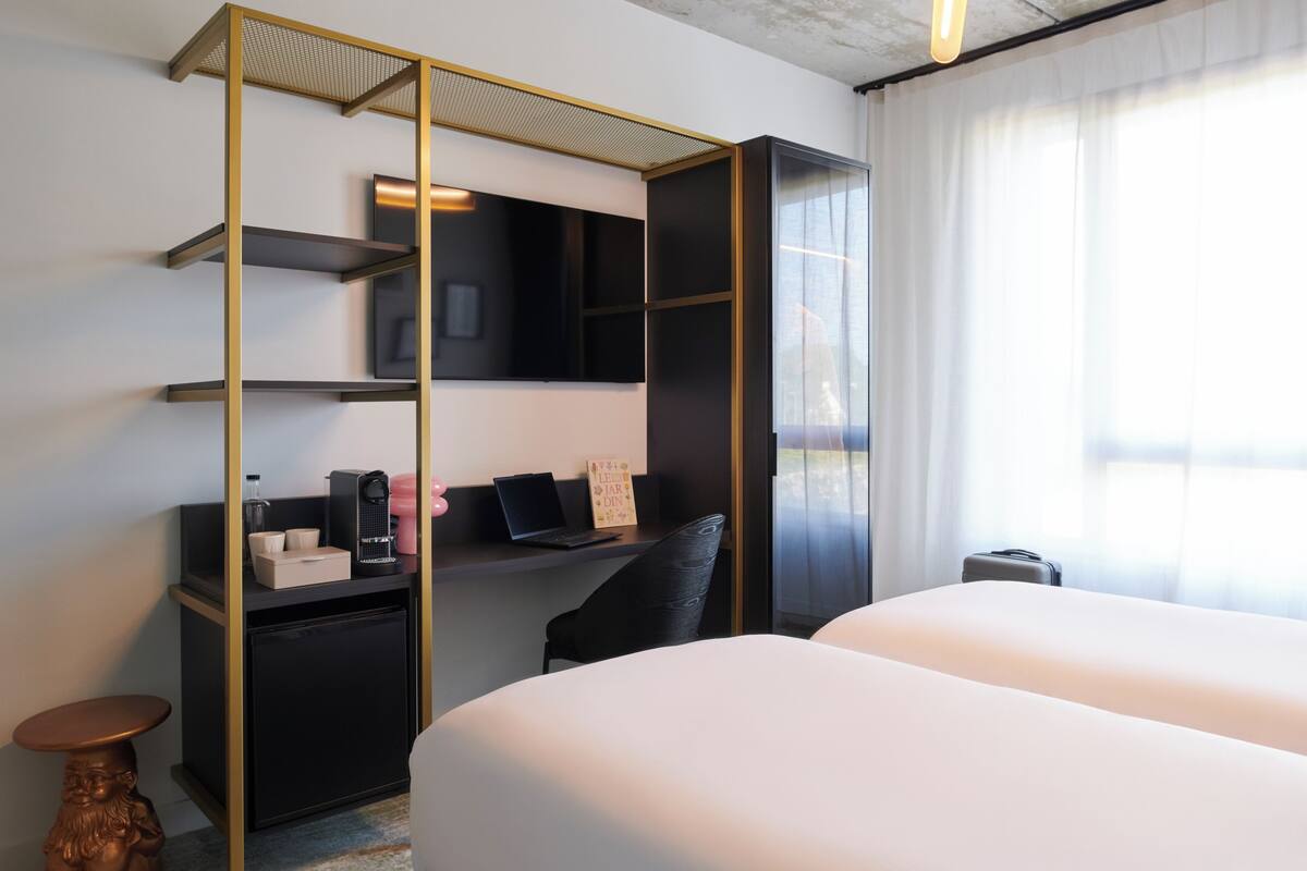 Habitación de hotel con cama, escritorio con computadora, refrigerador pequeño, televisión y ventana con cortinas.