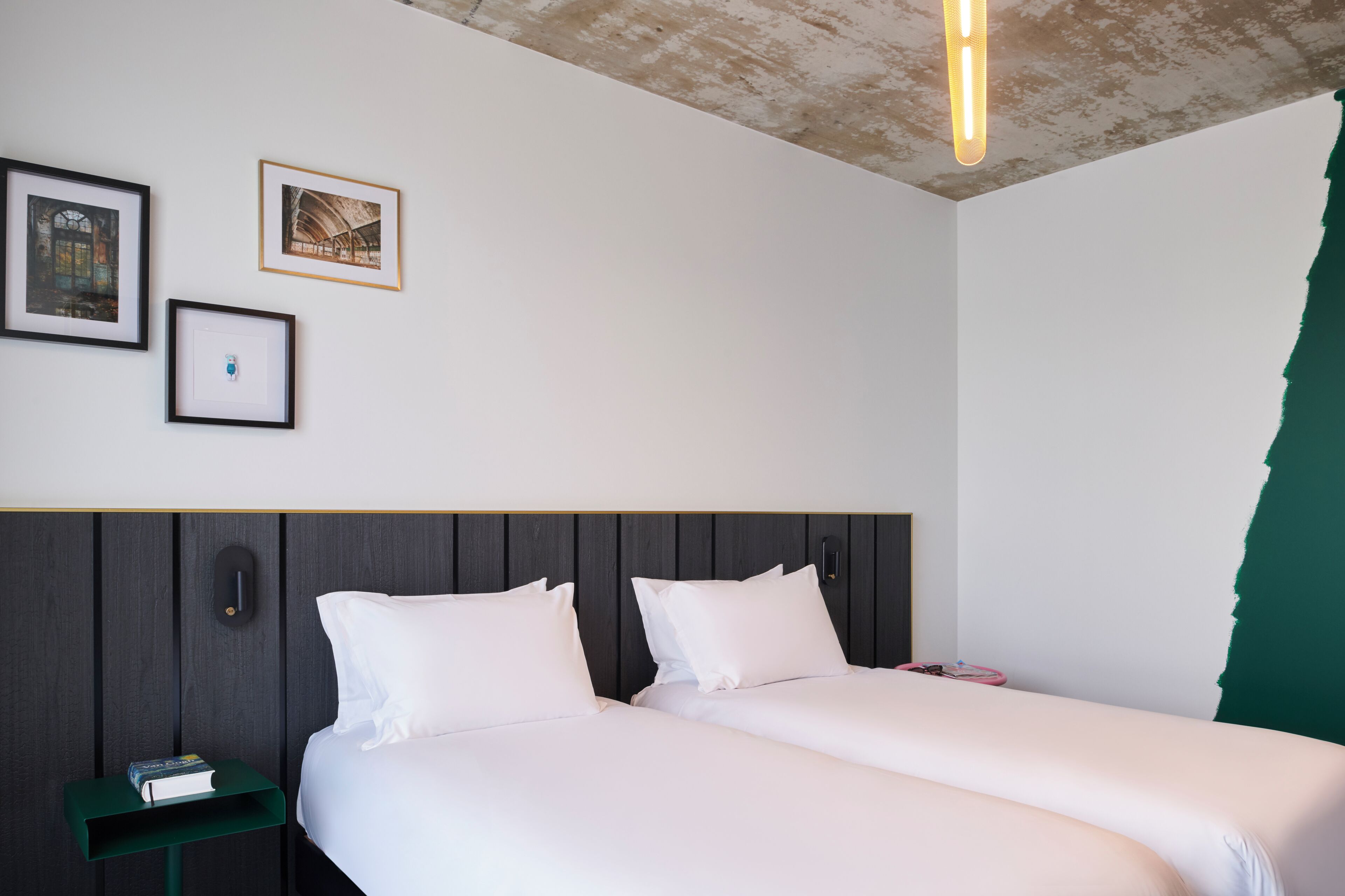 Habitación de hotel con dos camas, una mesita de noche con un libro, cuadros en la pared y una pared de acento verde.