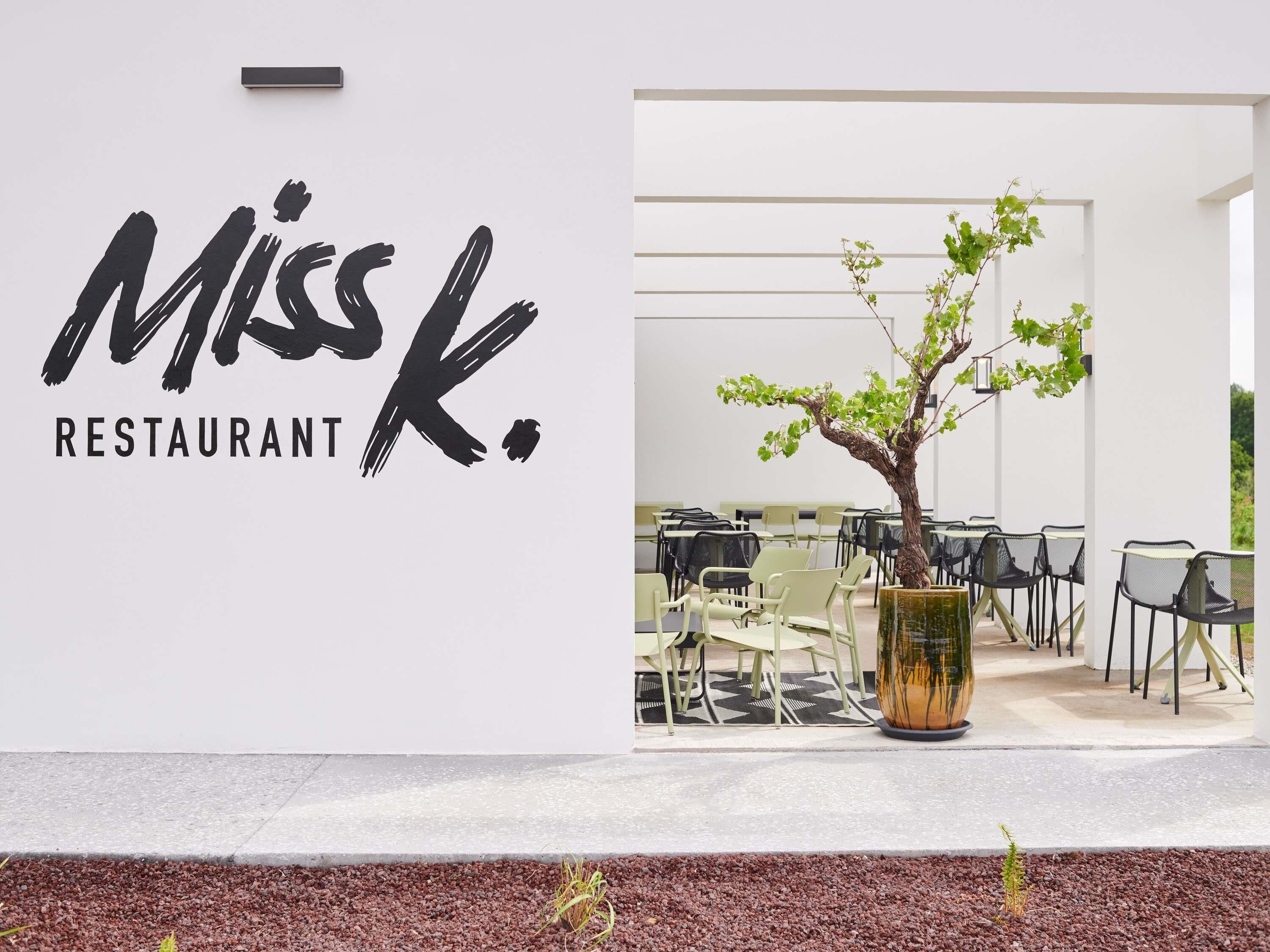Une enseigne de restaurant portant le nom « Miss K. » et un espace avec des tables et des chaises où les clients peuvent s’asseoir.