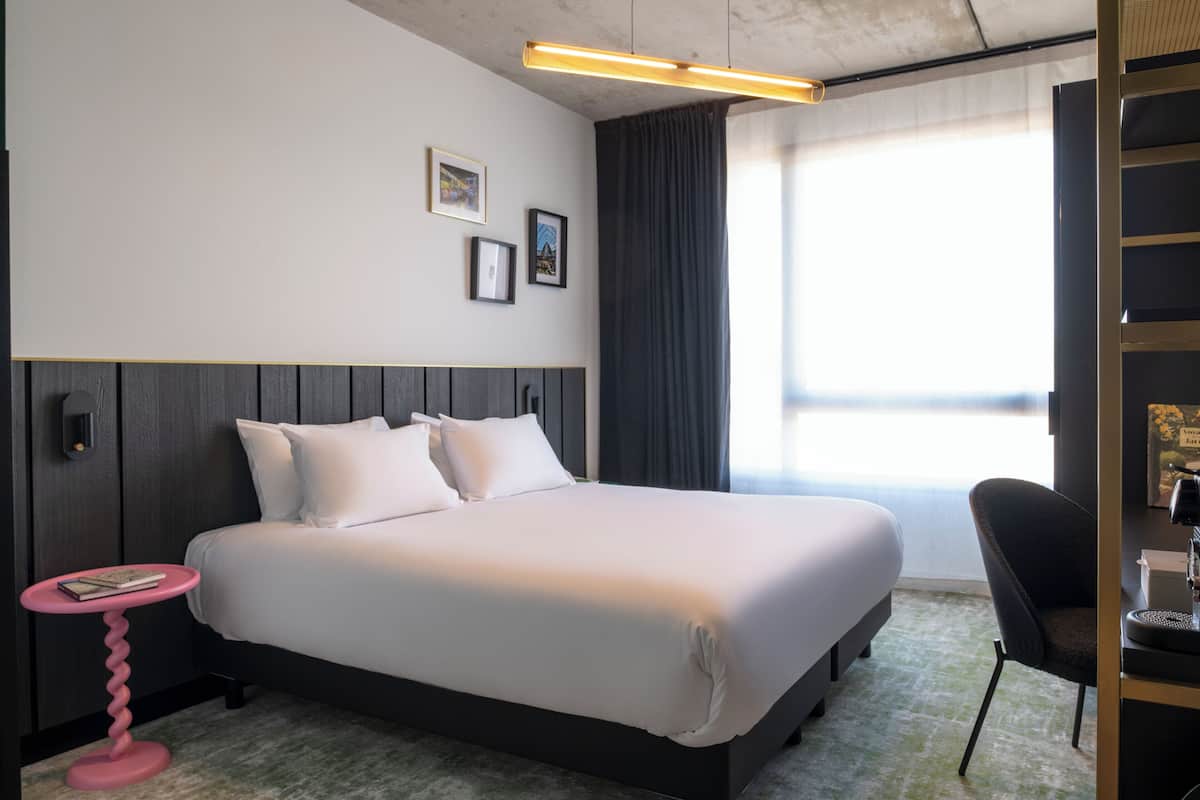 Una habitación de hotel con una cama, una mesita redonda pequeña, una silla, un escritorio con lámpara y una ventana con cortinas.