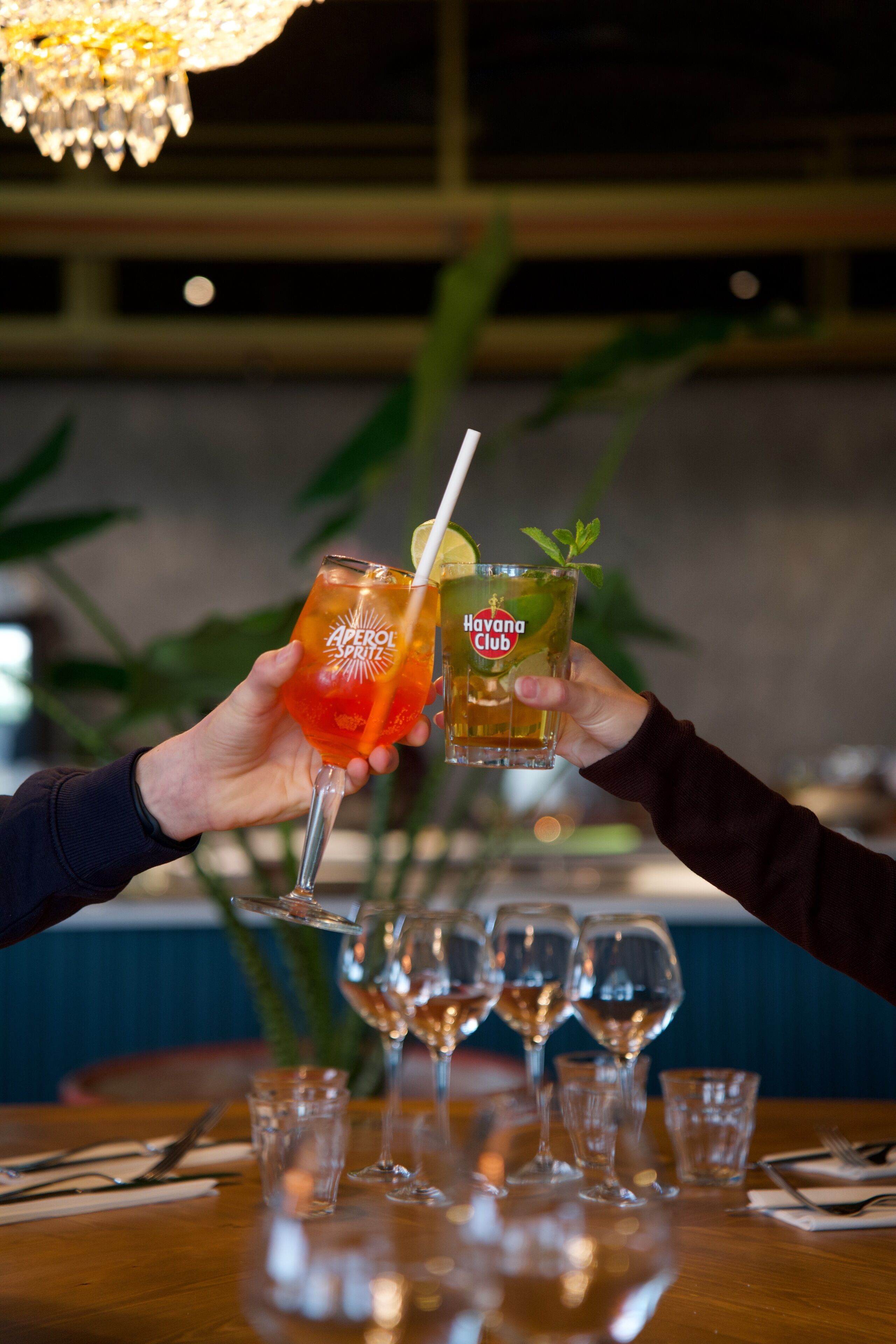 Deux mains tenant des verres ornés d’étiquettes « Aperol Spirit » et « Havana Club », une table en bois avec des verres à vin, et un lustre en arrière-plan.