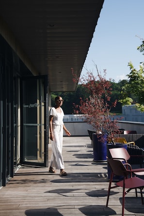 Une femme vêtue de blanc se promène sur une terrasse en bois, où se trouvent des meubles d’extérieur et une plante en pot.