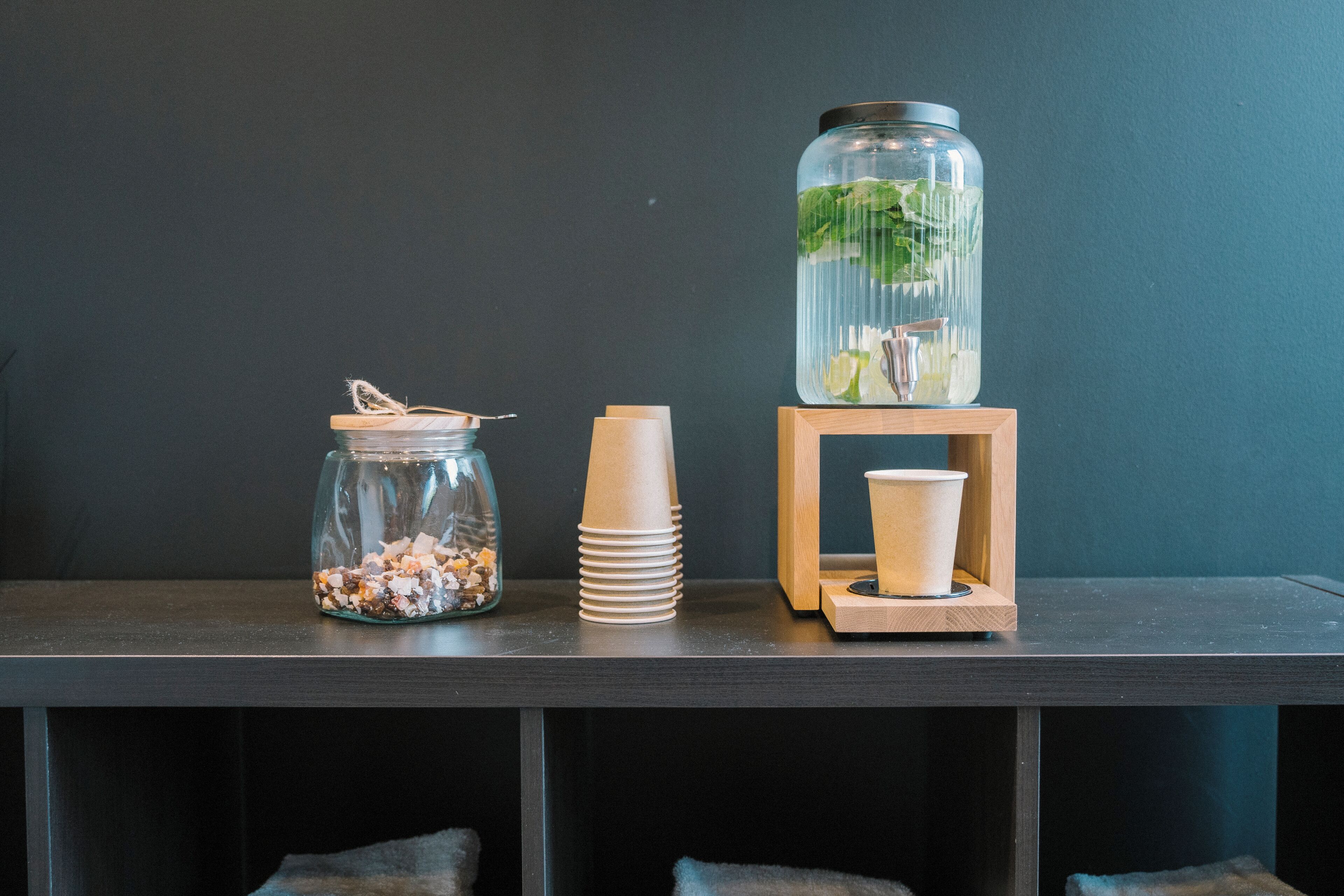Una repisa con un tarro de vidrio lleno de objetos coloridos, una pila de vasos de papel y un recipiente alto de vidrio con tapa y hojas de menta.