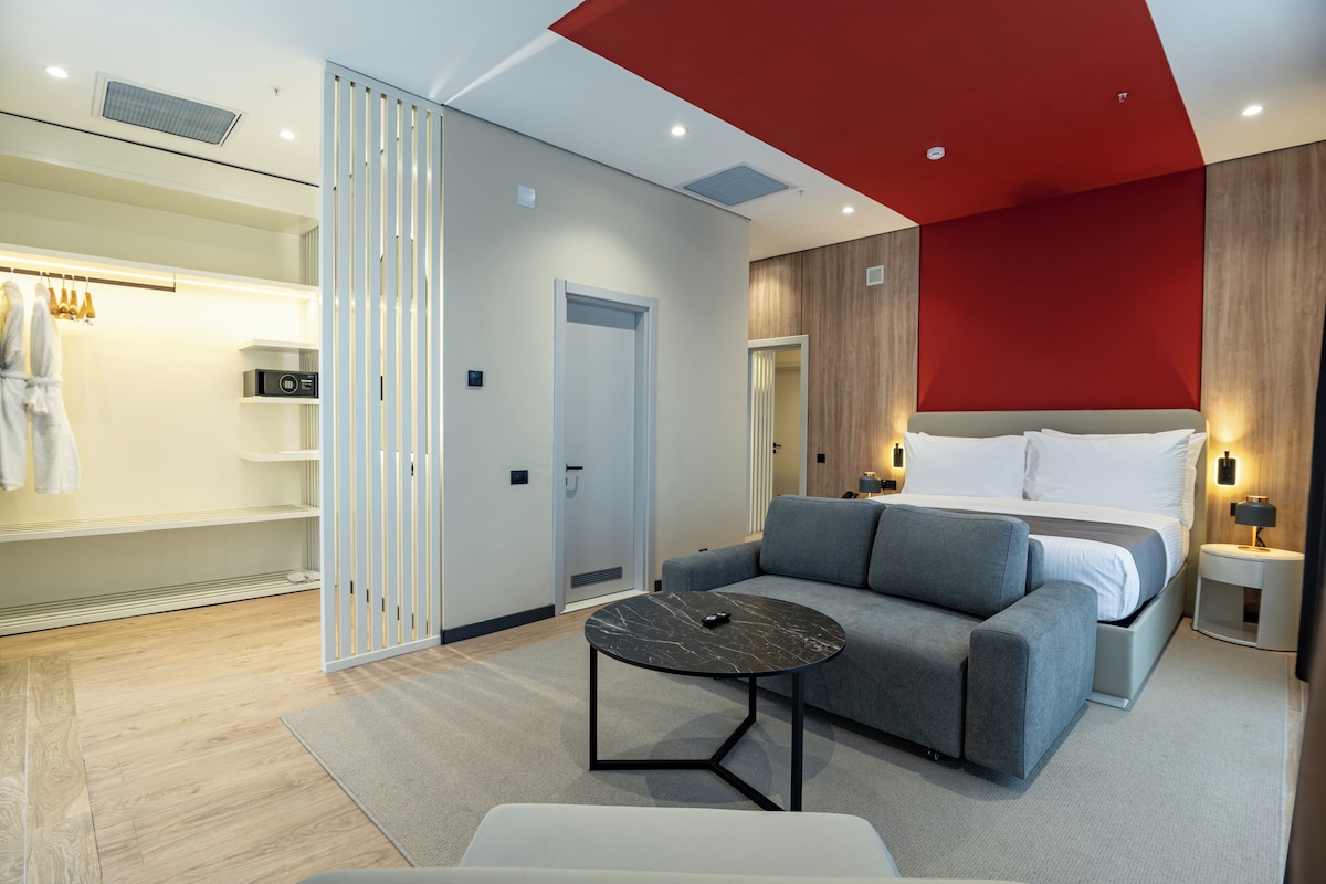A modern hotel room with a bed, a sofa, a small table, a wardrobe, and a red accent wall.
