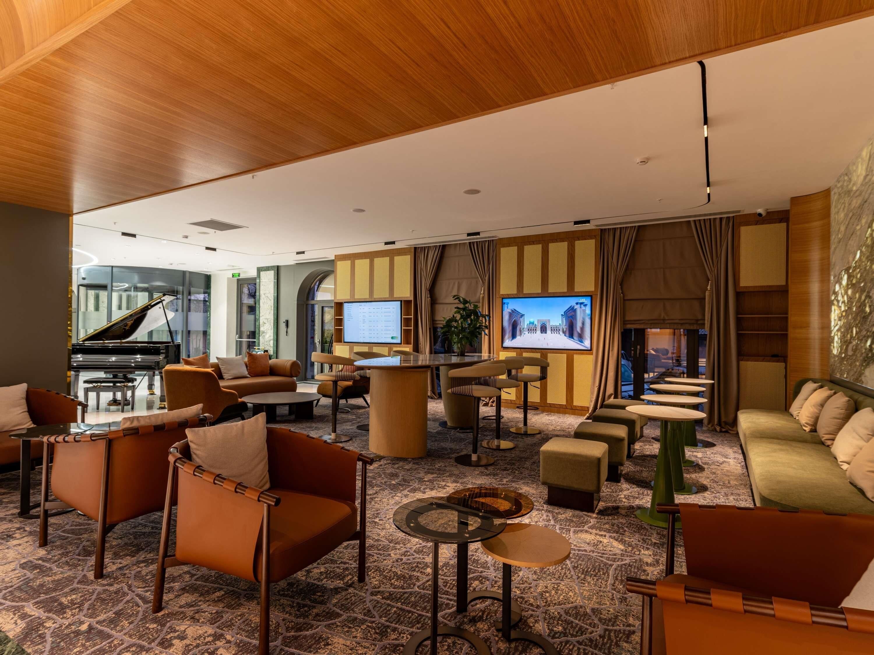 A spacious lobby with a grand piano, comfortable seating, a large TV screen, and a decorative wall.