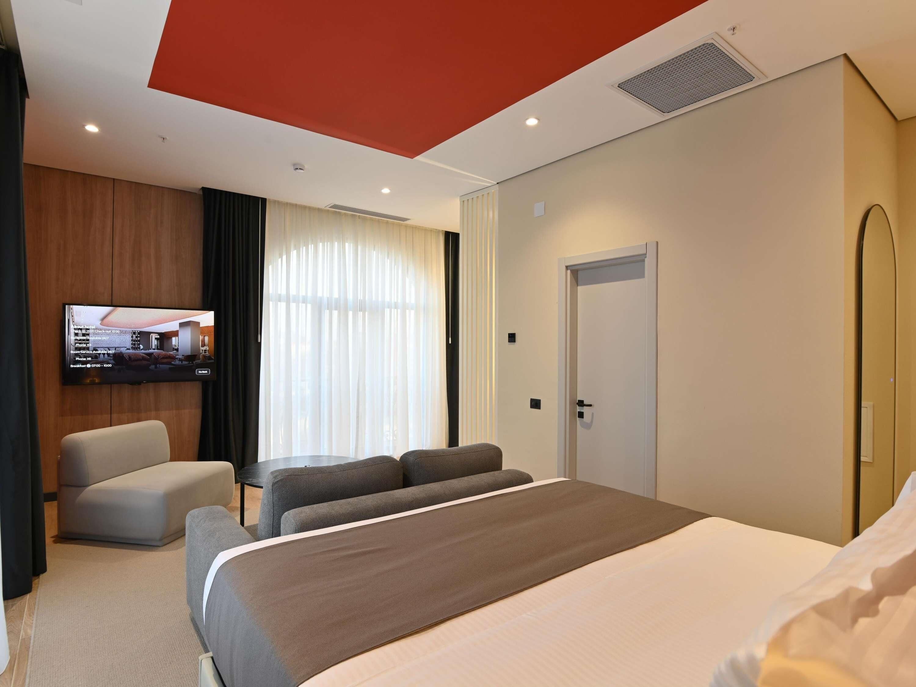 A hotel room with a bed, a TV mounted on the wall, a seating area with a sofa and armchairs, and a door.