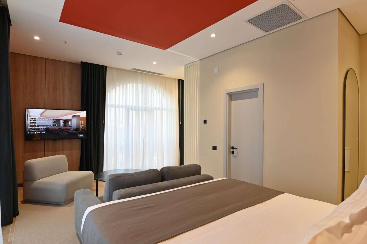 A hotel room with a bed, a TV mounted on the wall, a seating area with a sofa and armchairs, and a door.