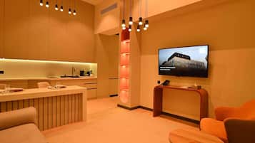 Un hall d’hôtel moderne doté d’un espace cuisine, d’un téléviseur affichant le nom de l’hôtel et de différents espaces pour s’asseoir.
