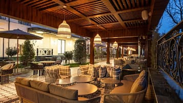 An outdoor seating area with wicker furniture and round tables, under a wooden ceiling with slatted beams.