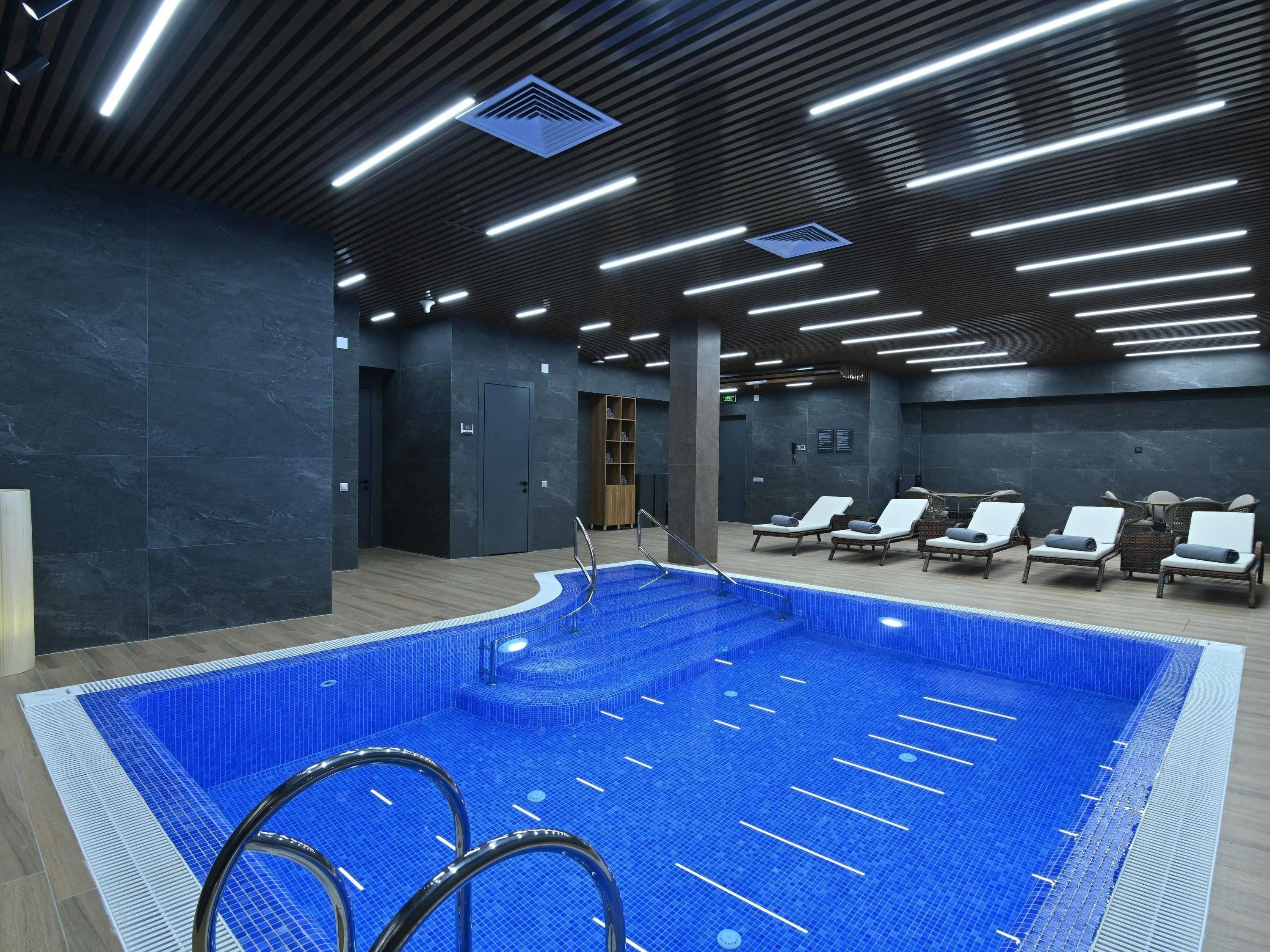 An indoor swimming pool with a tiled floor, surrounded by lounge chairs and a modern ceiling design with linear lighting.