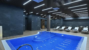 An indoor swimming pool with a tiled floor, surrounded by lounge chairs and a modern ceiling design with linear lighting.