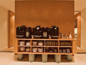 A coffee station with multiple espresso machines, a variety of cups and saucers, and a wooden panel backdrop. - Mercure Tashkent North (Tashkent)