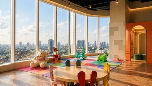 A high-rise room with a large window offering a city view, a round table with colorful chairs, and a teddy bear with a heart-shaped pillow. - Novotel Jakarta Pulo Mas (Jakarta)