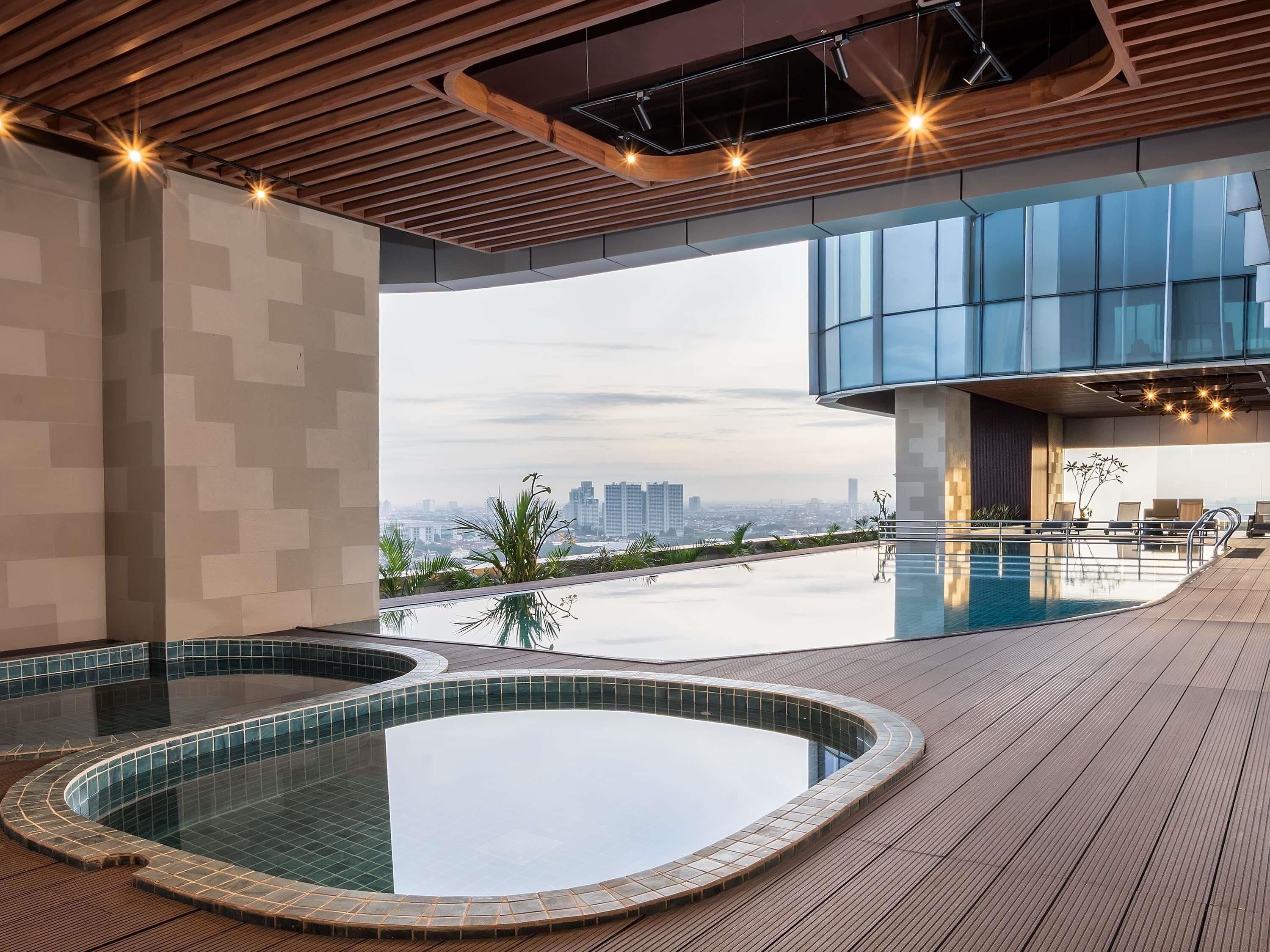 Una piscina interior moderna con vista a la ciudad, terraza de madera y zona de descanso.