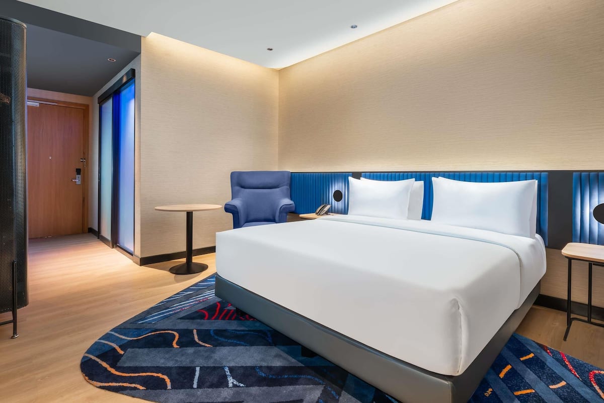 Una habitación de hotel moderna con una cama grande, una mesita redonda pequeña, un sillón azul y una puerta que da a otra habitación.
