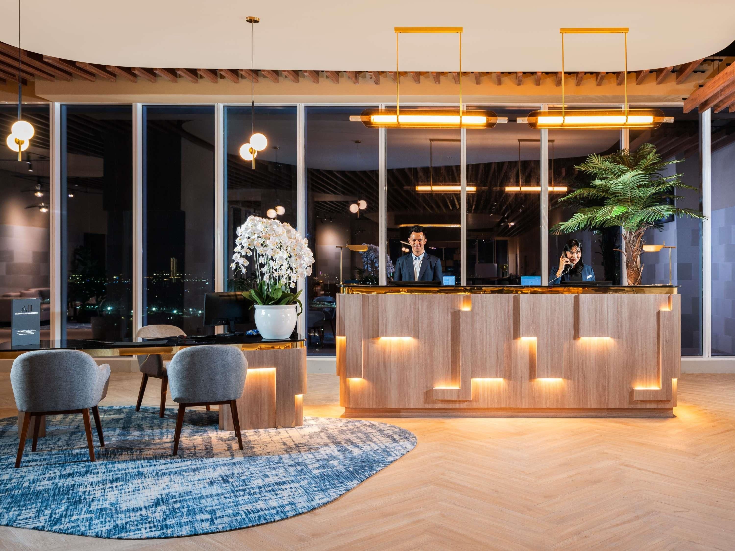 Un mostrador de recepción moderno con detalles de madera, un amplio ventanal con vista a la ciudad, y una sala de espera con dos sillas grises y un tapete azul estampado.
