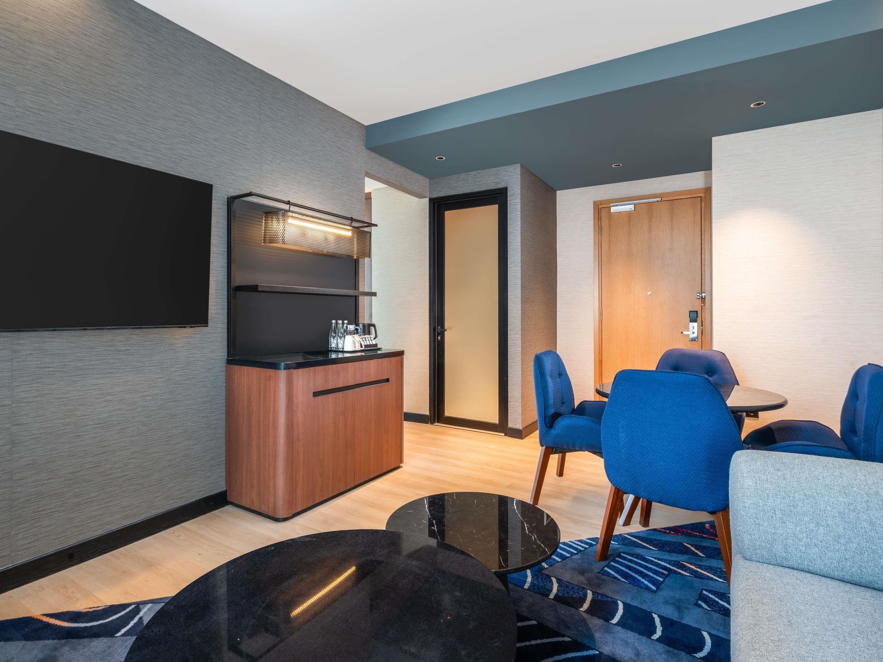 A modern hotel room with a kitchenette, a flat-screen TV, a dining area with blue chairs, and a wooden door.