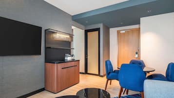 A modern hotel room with a kitchenette, a flat-screen TV, a dining area with blue chairs, and a wooden door.