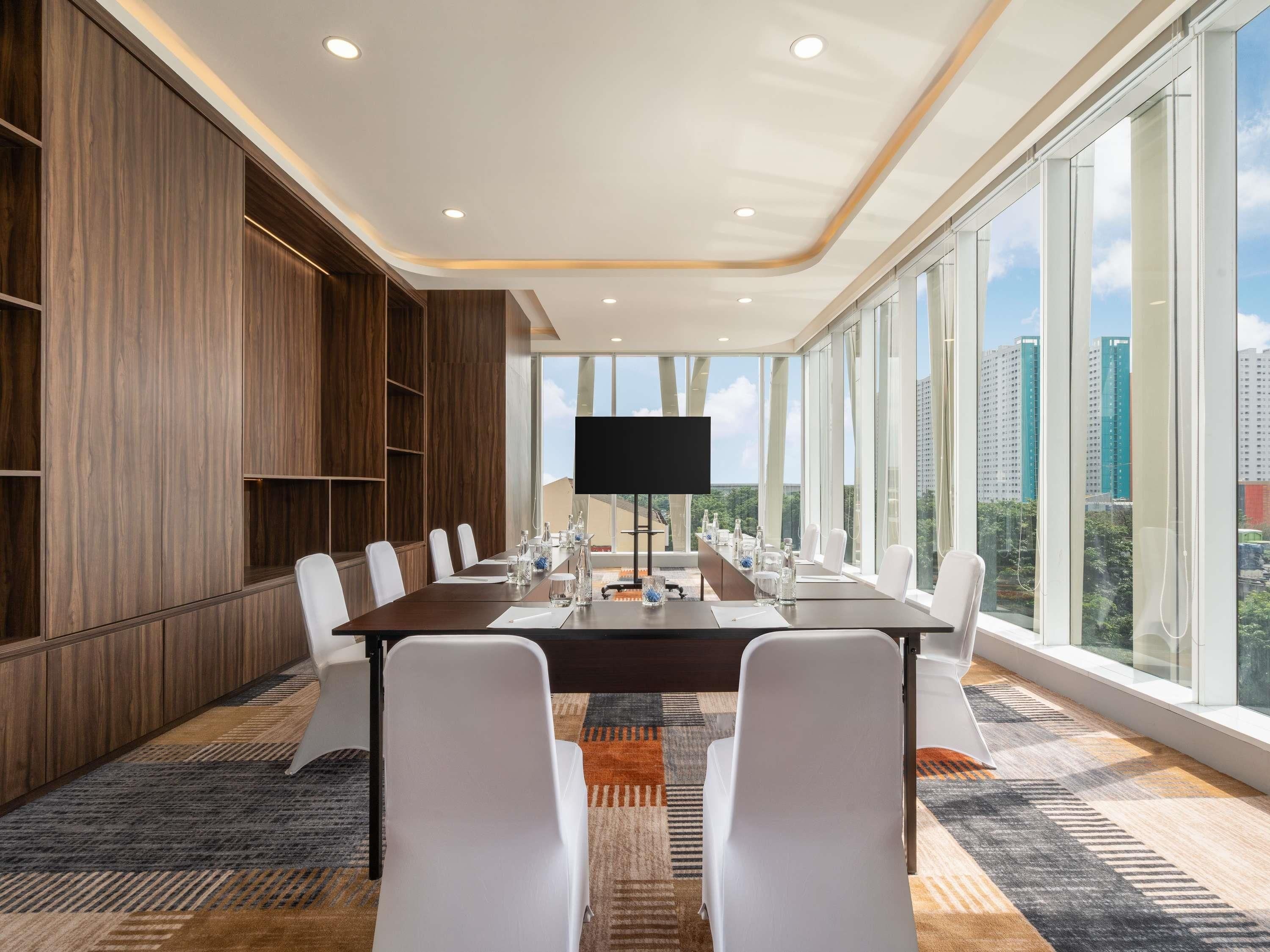A conference room with a long table, white chairs, a flat-screen TV, and large windows offering a city view.