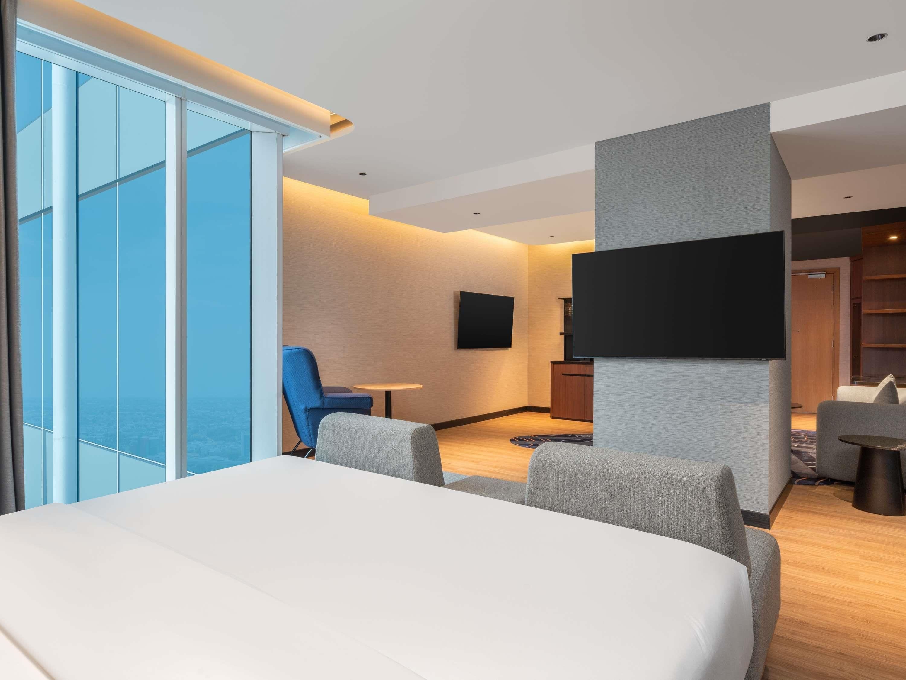 A hotel room with a bed, a TV mounted on the wall, a seating area with chairs, and a view through the window.