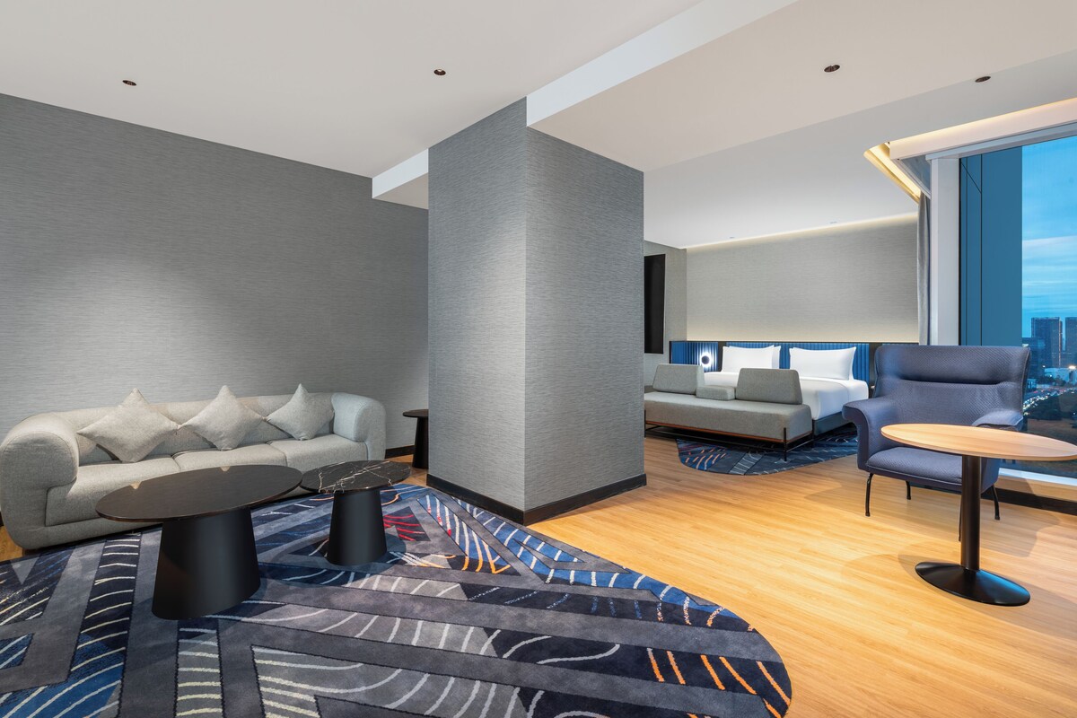Ein modernes Hotelzimmer mit grauem Sofa, einem runden Tisch und Blick auf die Stadt durch große Fenster.
