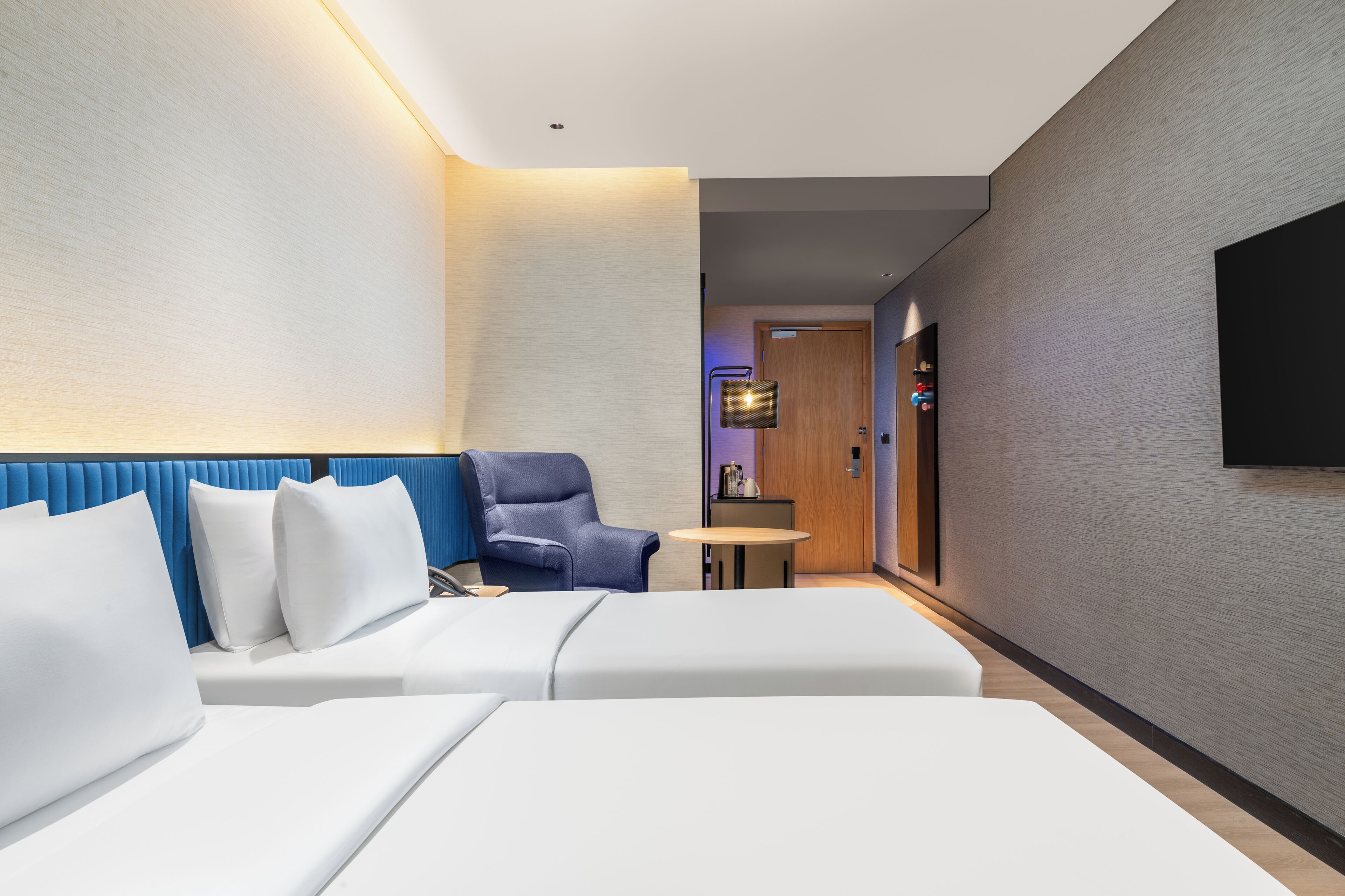 Una habitación de hotel moderna con una cama, un sillón azul, una mesita y un televisor montado en la pared.