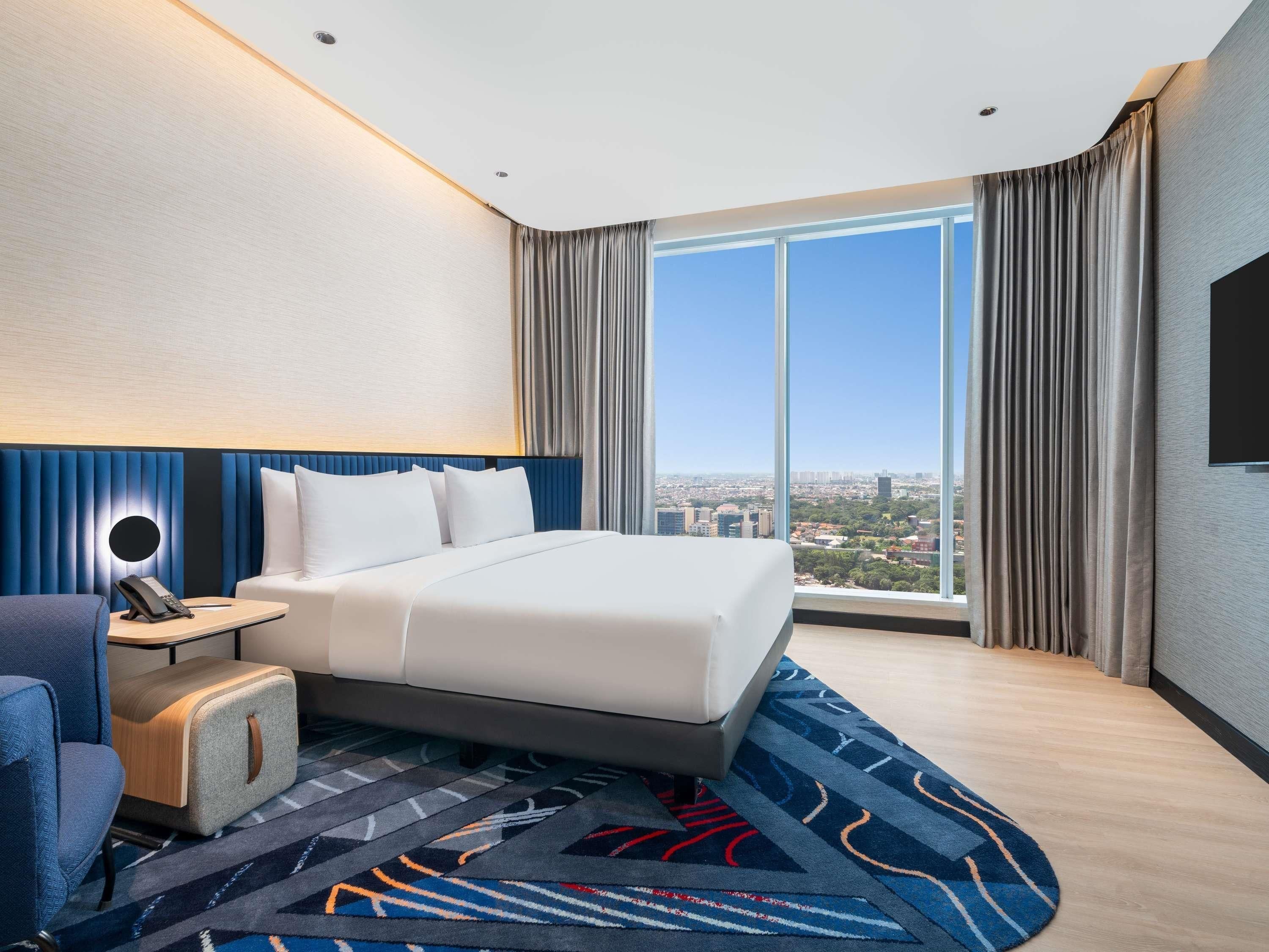 A hotel room with a large bed, a desk with a phone, a chair, a small ottoman, a patterned rug, and a city view through large windows.