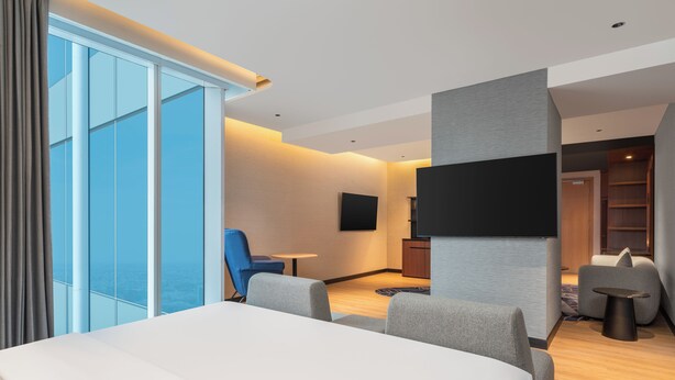 A modern hotel room with a large bed, a flat-screen TV, a seating area with a small table, and a view of the city through large windows. - Novotel Jakarta Pulo Mas (Jakarta)